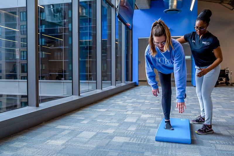 A physical therapist closely spots a woman wearing a blue Tufts Physical Therapy sweatshirt as she performs a single-leg balancing exercise on a blue foam pad in a brightly lit room with large floor-to-ceiling windows