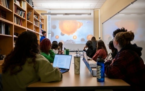 A group of students in a classroom watch a scientific video projected on a screen, with laptops and water bottles on the table.